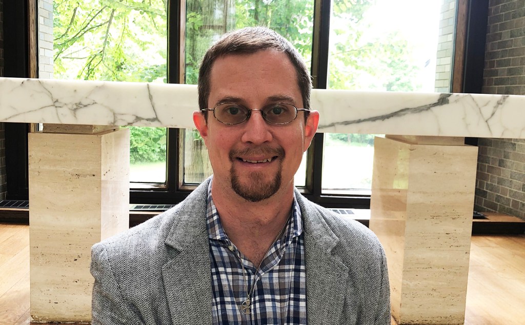Brother Larry Whitney seated, smiling in front of the altar in Brandeis University's Harlan Chapel trees and sunlight in the background.